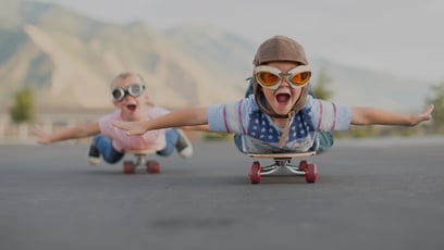 Zwei Kinder fahren auf Skateboards eine Straße hinunter. Sie liegen dabei auf dem Bauch. Beide haben Helm und Pilotenbrille auf. Sie träumen davon, zu fliegen.